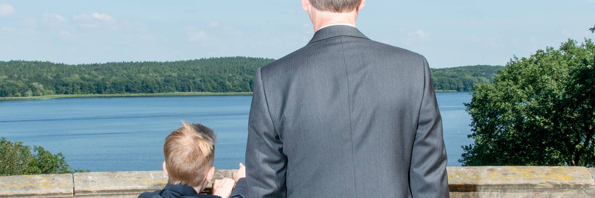 A man and his son, both in suits, look out over a body of water.