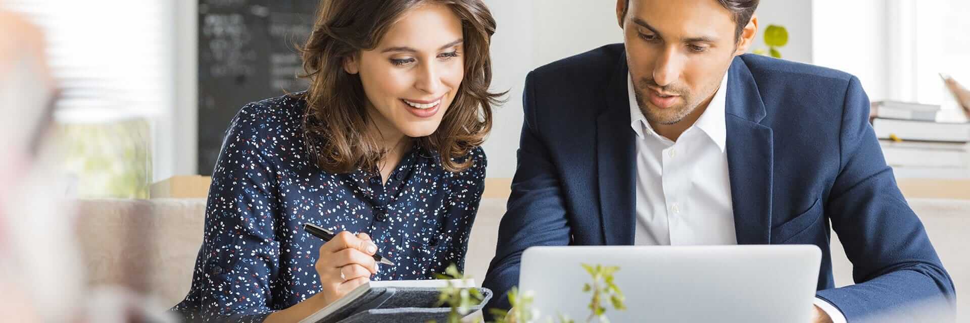 A male and female professional consulting over a laptop while taking notes.