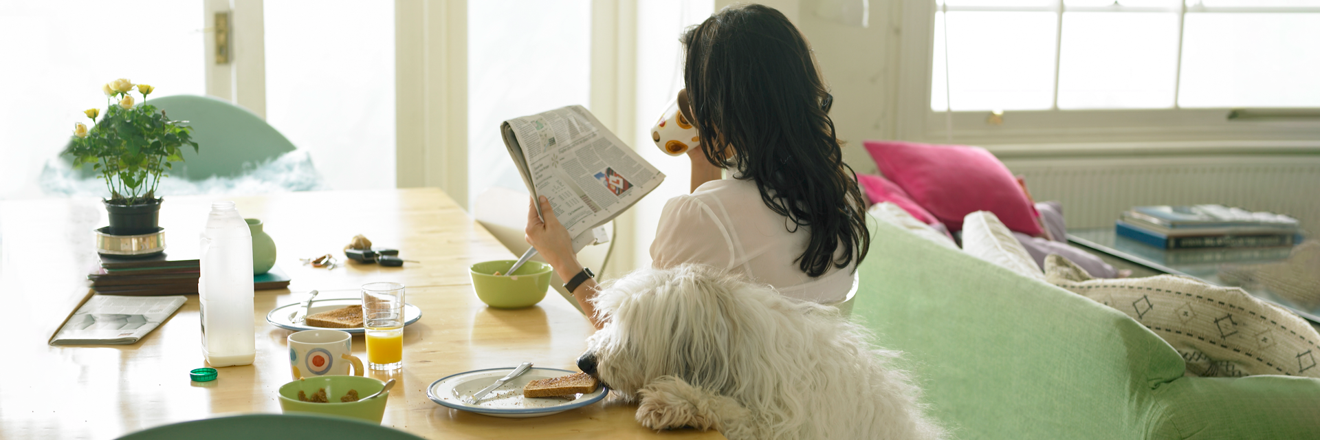 A woman sits at a dining room table, drinking coffee and reading a newspaper.