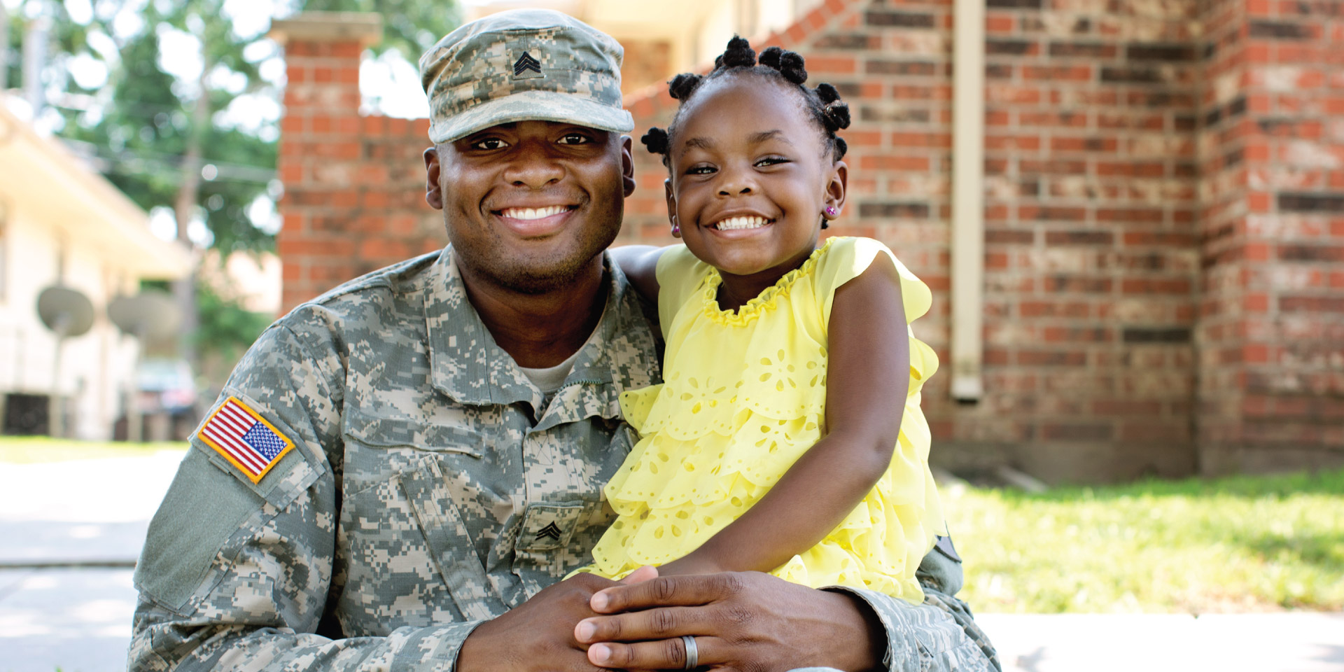 A man in a military uniform squats and smiles with his daughter on his knee.