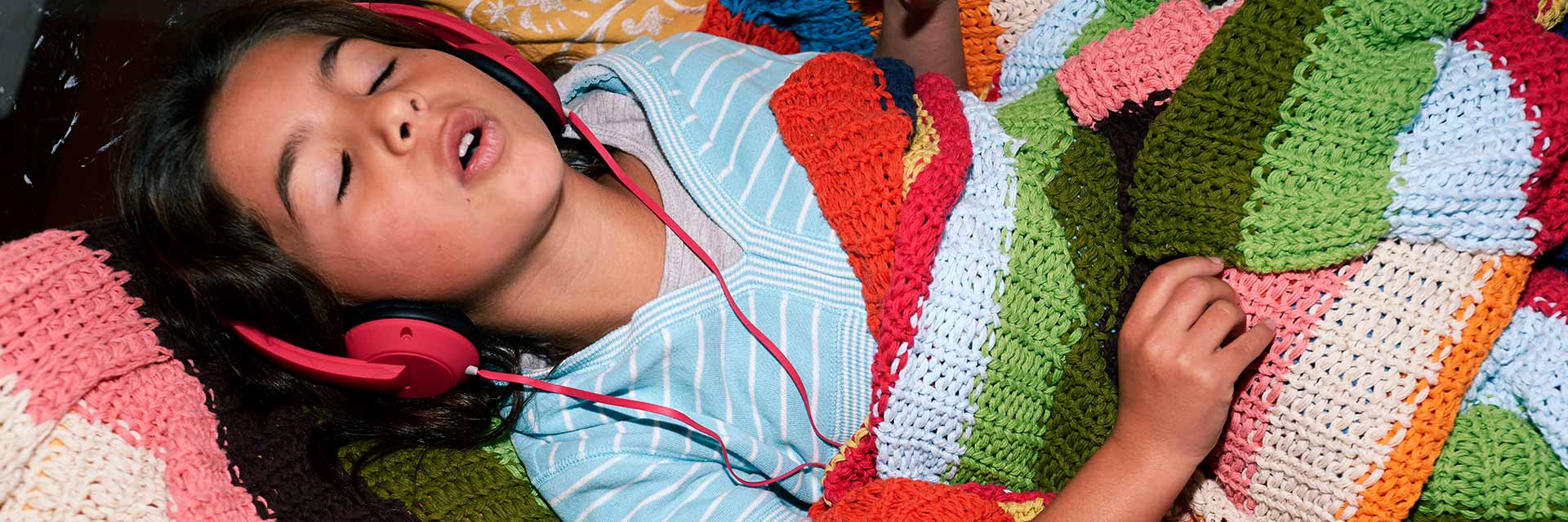 A young girl listens to music through headphones while snuggling with a blanket.